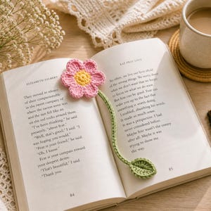 Puede incluir: Un marcapáginas de ganchillo con forma de flor rosa y amarilla con tallo y hoja verdes, colocado en un libro abierto. Las páginas del libro muestran texto de "Eat Pray Love" de Elizabeth Gilbert. Una taza de café está cerca.
