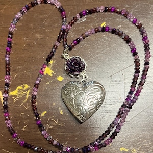 May include: A necklace featuring a long strand of small, faceted beads in varying shades of purple and pink. The pendant consists of a dark purple rose-shaped charm attached to a silver-toned heart-shaped locket with an embossed floral pattern.
