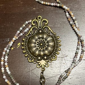 May include: A necklace featuring a large, ornate, antique brass-toned pendant with a central floral motif and scrollwork details. A small, clear, faceted bead hangs from the bottom. The chain consists of multi-colored, small, faceted glass beads.