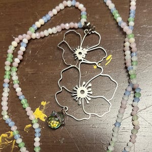 May include: A long necklace featuring a strand of small, faceted pastel-colored beads in shades of pink, blue, and green. A large, silver-toned metal pendant shaped like two overlapping flower outlines hangs from the beaded chain with a small, iridescent, faceted glass bead charm attached at the bottom.