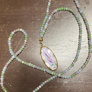 May include: A long beaded necklace featuring small, faceted glass beads in alternating shades of pale green, soft lavender, and translucent white. The necklace includes a central oval pendant with a gold-toned metal frame and a lavender floral design.