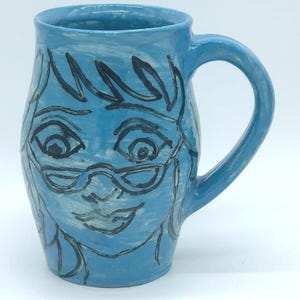 May include: A light blue ceramic mug with a whimsical face design. The face is outlined in black, featuring eyes, glasses, and a smiling mouth. The mug has a curved handle and is set against a white background.