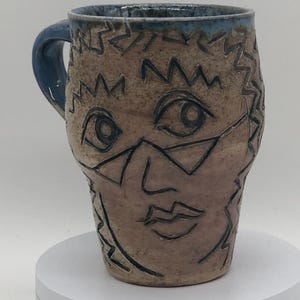 May include: A handmade ceramic mug with a face design. The mug features a tan base color with a blue rim and handle. The face is outlined in black, with glasses and a mouth. The mug is sitting on a white platform.