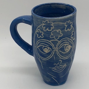 May include: A blue ceramic mug with a handle, featuring a white sketched face design with glasses and floral accents. The mug is a cylindrical shape and sits on a white surface.