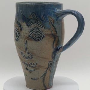 May include: A tall ceramic mug with a face design. The mug features a blue handle and rim, with a tan and brown body. The face is etched into the mug, with blue accents. The mug is on a white surface.