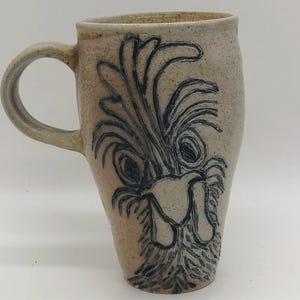 May include: A handmade ceramic mug with a tan speckled finish and a dark blue rooster design. The mug has a curved handle and a cylindrical shape, perfect for coffee or tea.