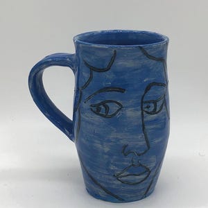 May include: A blue ceramic mug with a handle, featuring a hand-drawn face design in black. The mug has a textured surface and a simple, artistic aesthetic. The mug is suitable for hot or cold beverages.