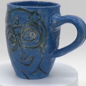 May include: Blue ceramic mug with a face design. The mug features a blue glaze with a face design in black and gold. The mug has a curved handle and is sitting on a white surface.