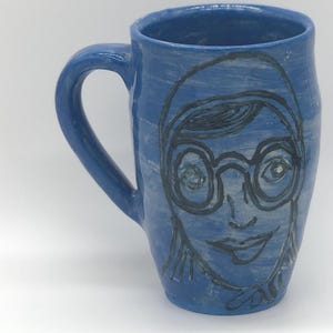 May include: A white ceramic mug with a black handle and the text "But First, Coffee" in a black, handwritten-style font. The mug is on a white surface.