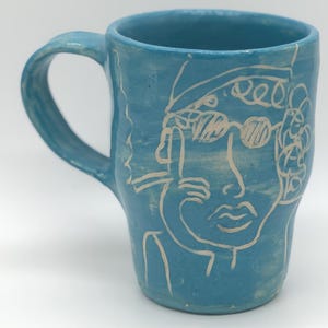 May include: A white ceramic mug with a black handle and the text "But First, Coffee" in a black, handwritten-style font. The mug is on a white surface.
