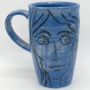 May include: A blue ceramic mug with a handle, featuring a hand-drawn face design. The face is outlined in black, with details like eyes, a nose, and a mouth. The mug has a smooth, slightly textured surface.