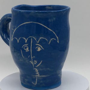 May include: A cobalt blue ceramic mug with a handle. The mug features a white line drawing of a face partially covered by an umbrella. The mug is sitting on a white surface.