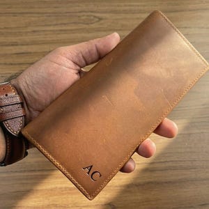 Könnte beinhalten: Eine braune Ledergeldbörse mit den Initialen "AC" auf der unteren rechten Ecke. Die Geldbörse ist rechteckig und scheint aus hochwertigem Leder gefertigt zu sein. Eine braune Lederuhr mit silberner Schnalle ist im Bild zu sehen.