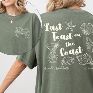 May include: Moss green t-shirt with an ivory design. The front features two champagne glasses with bows. The back displays the text "Last Toast on the Coast" with seashell and cocktail graphics. "Amanda's Bachelorette" and "est. 2026" are also printed.