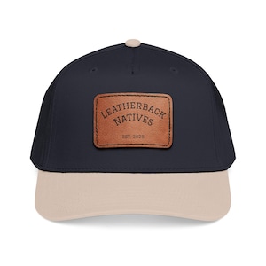 May include: A navy blue baseball cap with a beige peak and a brown leather patch. The patch reads "LEATHERBACK NATIVES" above "EST. 2025". The cap has a curved peak and a button on top.
