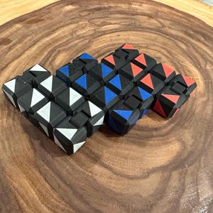 May include: A colorful puzzle toy with a snake-like design. The puzzle features black cubes with white, blue, and red triangular accents. The toy is displayed on a wooden surface.