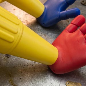 May include: A close-up of three colorful hand-shaped objects. A red hand is positioned with a yellow tube-like object inserted into the palm. A blue hand is in the background. The objects are on a metallic surface with small mechanical parts.