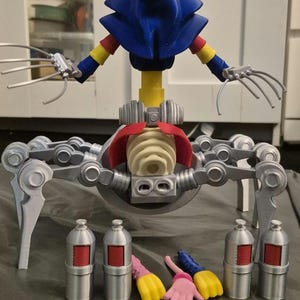 May include: A whimsical, articulated toy figure with a jester-like head in blue, red, and yellow. The figure has a skeletal torso, silver mechanical legs, and claw-like hands. Accessories include silver canisters and colorful hand-shaped pieces.