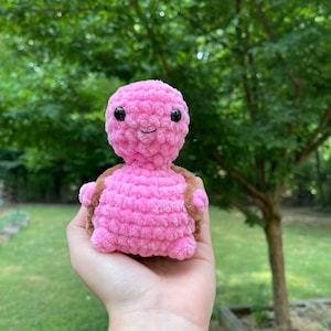 May include: A handmade pink crochet turtle with a smiling face and black eyes. The turtle has a brown shell and is held in a hand against a blurred green background. The plush toy is soft and cuddly.