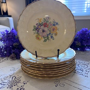 May include: A stack of cream-colored vintage plates with a gold rim and a floral design in the center. The plates have a scalloped edge and are stacked on a white tablecloth.