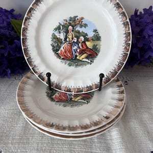 May include: A set of three white ceramic plates with a scalloped edge and a metallic gold-toned trim. The center of each plate features a colorful illustration of a couple in historical attire in a garden setting with trees and a stone structure.