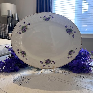 May include: An oval, white porcelain serving platter with a gold rim and a basketweave texture. The platter is decorated with clusters of purple flowers. The platter is displayed on a gold stand and surrounded by purple flowers.