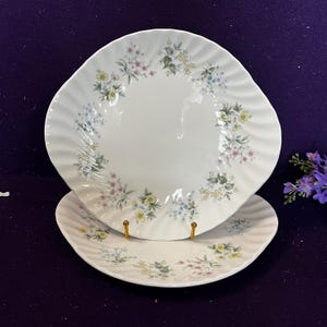 May include: Two white ceramic plates with a scalloped, wavy edge design. The plates feature a delicate floral pattern around the rim, consisting of small yellow, pink, and blue flowers with green leaves arranged in a circular wreath motif.