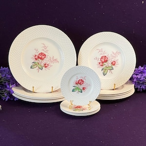 May include: A set of white ceramic plates featuring a textured basket-weave pattern on the rim and a central floral motif of red roses with green leaves. The collection includes multiple sizes of round plates stacked in three distinct groupings.