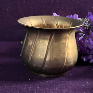 Pode incluir: Uma pequena taça de metal em tom dourado com uma borda alargada e uma série de reentrâncias verticais e diagonais ao longo do seu corpo arredondado. A superfície tem um acabamento polido e reflexivo que capta a luz com suavidade.