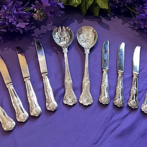 May include: A set of eight silver-toned metal utensils including four knives on the left, a salad fork and spoon in the centre, and three additional knives on the right. Each piece features an ornate, embossed floral pattern on the handle.