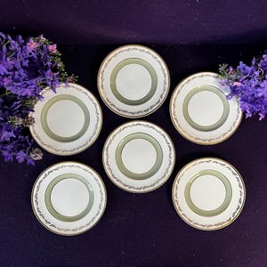 Può includere: Un set di cinque piatti rotondi in ceramica con centro bianco, una fascia verde salvia e un bordo dorato decorato con un motivo a foglie ripetuto. I piatti sono disposti in gruppo su una superficie di colore viola scuro.