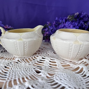 May include: A cream-coloured ceramic creamer and sugar bowl set. The creamer has a handle and spout, while the sugar bowl is round. Both pieces feature a textured design with a yellow interior, set on a white lace doily.
