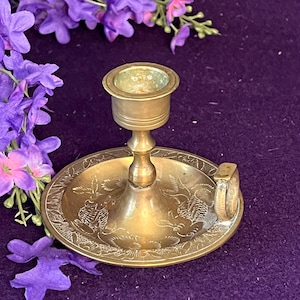 Puede incluir: Un candelabro de latón con base circular que presenta un patrón floral grabado y una pequeña asa lateral. El candelabro tiene un soporte cilíndrico central para una vela y un acabado metálico pulido.