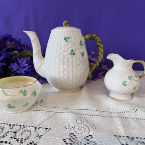 Puede incluir: Un juego de té de cerámica blanca con textura tejida, con detalles de tréboles verdes. El juego incluye una tetera, una jarra de crema y un azucarero. La tetera tiene un asa y un pico tejidos con detalles amarillos.