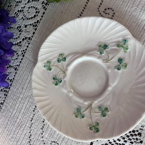 Puede incluir: Platillo de cerámica blanca con borde festoneado y diseño espiral en relieve. Presenta detalles de tréboles verdes. Una pieza delicada para servir té o exhibir objetos pequeños. El platillo mide aproximadamente 13 cm de diámetro.