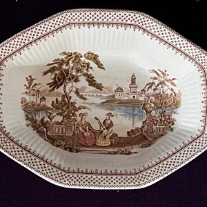 May include: An octagonal ceramic bowl with a cream-coloured base and a brown transferware pattern. The rim features a repeating geometric border. The centre depicts a landscape with a lake, buildings, trees, and two figures sitting on a stone terrace.