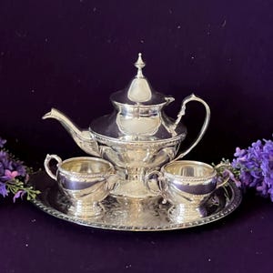 May include: A polished silver tea set comprising a teapot, a creamer, and a sugar bowl set upon an oval serving tray. Every item displays a decorative rope-style border detail. The teapot features a pointed lid finial and a curved handle.