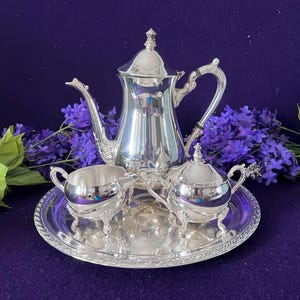 Puede incluir: Un juego de té plateado que consta de una tetera alta con tapa, una jarra para crema y un azucarero con tapa, todo dispuesto sobre una bandeja de servicio redonda de plata pulida con un borde decorativo en relieve y asas curvas.