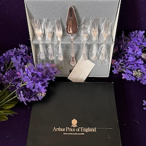 Peut inclure: Un ensemble de service à gâteau argenté Arthur Price of England, comprenant une pelle à tarte et six fourchettes à pâtisserie. Les couverts présentent des manches ornés et sont présentés dans une boîte grise avec un couvercle noir marqué.