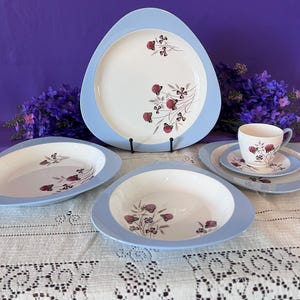 May include: A vintage porcelain dinnerware set with a light blue trim and floral design. The set includes a large serving plate, oval serving dishes, bowls, a teacup, and saucers. The floral pattern features red and purple flowers.