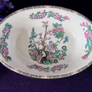 May include: An oval ceramic serving bowl with a cream-coloured base. The interior features a central illustration of a tree with pink flowers and green leaves, surrounded by floral motifs. The rim has a geometric brown and white border pattern.