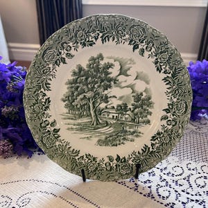 May include: A white and green decorative plate with a landscape scene in the center. The plate has a scalloped edge and a floral border. The landscape scene depicts a tree, a house, and a path. The plate is on a black stand.