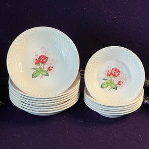 May include: A set of white ceramic bowls and plates featuring a textured lattice pattern on the rims and a central floral motif of red roses with green leaves and delicate pink sprigs. The collection includes two stacks of matching dinnerware.