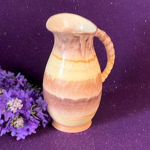 May include: A ceramic jug featuring a rounded body and a textured, rope-like handle. The exterior displays horizontal bands of cream, light orange, and muted brown colours with a drip-glaze effect near the rim and base of the vessel.