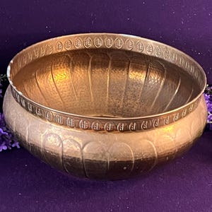 May include: A round, metallic, copper-toned bowl featuring a hammered texture on its exterior and interior surfaces. The upper rim is decorated with a repeating embossed leaf pattern, while the body displays vertical fluted indentations.