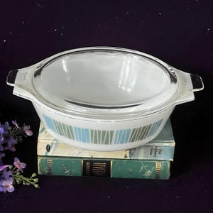 May include: A white glass casserole dish with a clear glass lid and integrated handles. The exterior features a repeating pattern of vertical light blue and sage green stripes. The dish has a smooth, rounded shape and a polished, reflective finish.