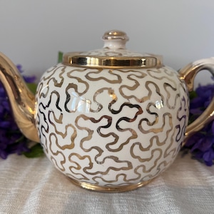 Puede incluir: Una tetera de cerámica blanca con un patrón dorado y detalles dorados en el pico, el asa y la tapa. La tetera tiene forma redondeada y está decorada con un diseño dorado en forma de garabatos. El fondo presenta flores moradas.