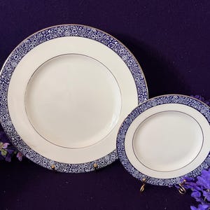 May include: Two round white ceramic plates featuring a wide border with a detailed dark blue and white floral pattern. Each plate has a thin gold rim along the edge of the centre basin and the outer circumference, creating a refined appearance.