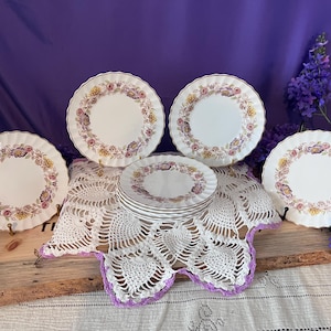May include: A set of white ceramic plates featuring a scalloped edge and a decorative border of colourful floral patterns in shades of pink, yellow, and purple. The collection includes four upright plates and a stack of smaller plates in the centre.