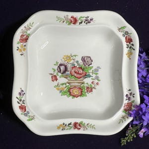 May include: A square white ceramic bowl with rounded corners, featuring a central floral basket illustration and smaller floral sprigs along the rim. The design includes red, purple, yellow, and green colors on a smooth, glossy white surface.